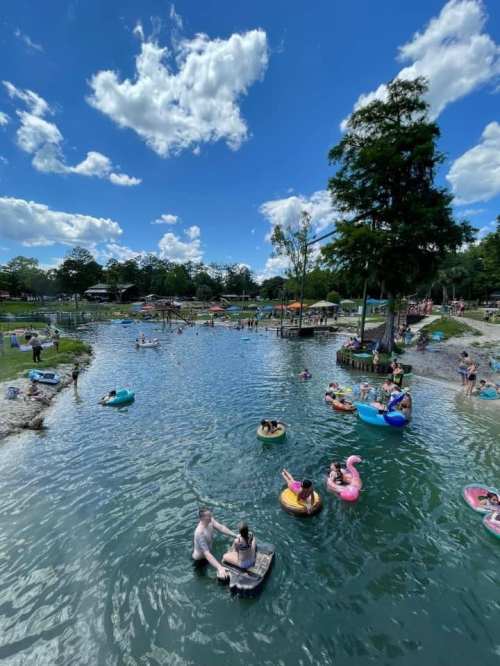 A sunny day at a recreational lake with people swimming and floating on colorful inflatables. Trees and clouds in the background.