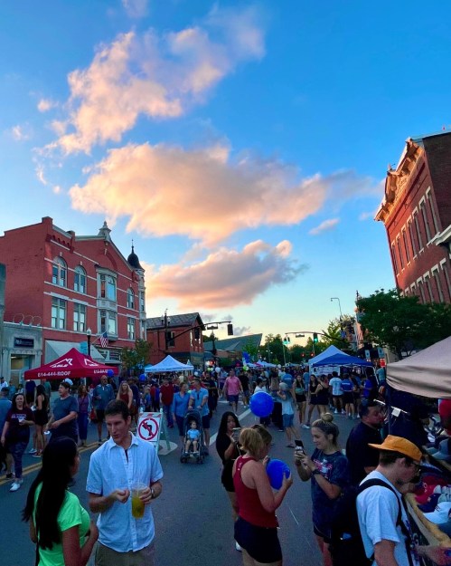 A lively street festival with crowds, tents, and colorful balloons under a blue sky with fluffy clouds.