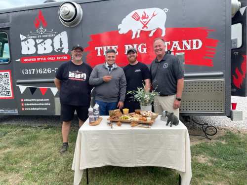 Four men stand in front of a BBQ food truck, smiling beside a table of food and decorations.