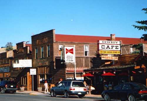 A rustic street scene featuring wooden buildings, a café sign, and parked cars under a clear blue sky.