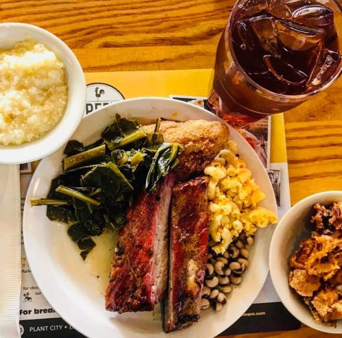 A plate of barbecue ribs, collard greens, macaroni and cheese, black-eyed peas, and cornbread, with a drink on the side.