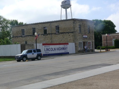 A stone building with a sign for the Lincoln Highway, parked vehicle, and American flags in a small town setting.