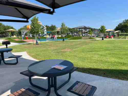 A sunny park scene featuring picnic tables, green grass, and playground equipment in the background.