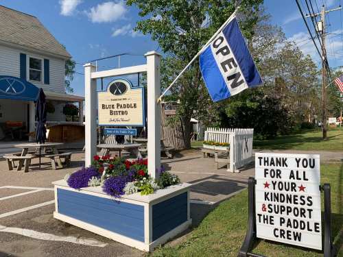 Exterior of Blue Paddle Bistro with a flag reading "OPEN" and a sign thanking customers for their support. Flowers in a planter.