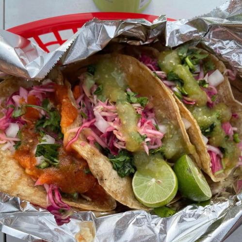 Three tacos in a basket, topped with colorful slaw, green salsa, and lime wedges, wrapped in foil.