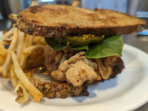 A crispy fried chicken sandwich with greens and mustard on seeded bread, served with a side of French fries.