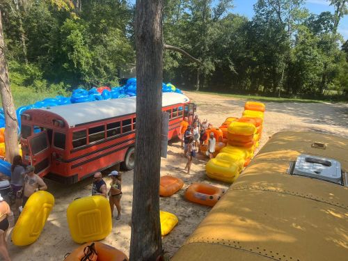 A red bus and yellow bus are surrounded by people and colorful inflatable rafts in a wooded area.