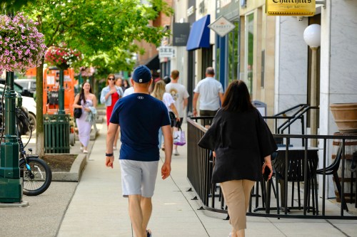 A couple walks down a busy sidewalk lined with shops and greenery on a sunny day.