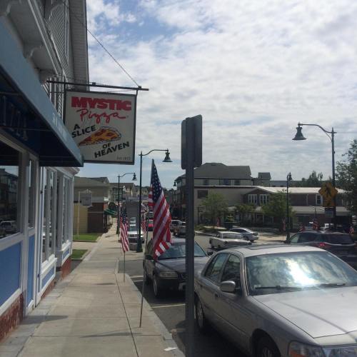 A street view of Mystic Pizza with American flags and a cloudy sky in the background.