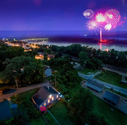 Aerial view of a river town at night, with colorful fireworks above and illuminated buildings along the water.