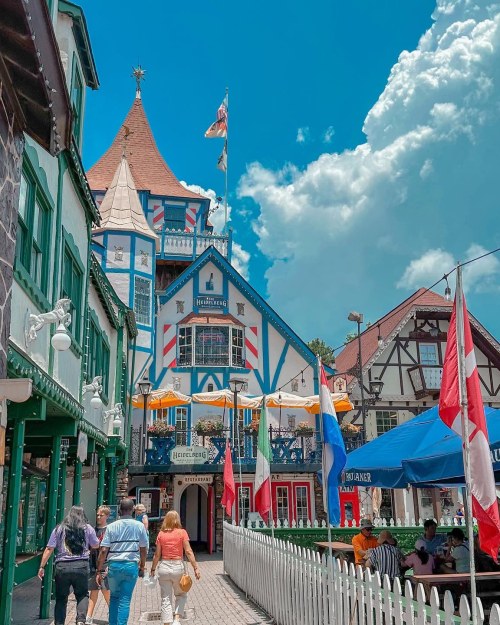 Colorful buildings with flags, people walking in a charming village under a bright blue sky with fluffy clouds.