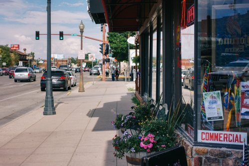 A sidewalk lined with flowers, storefronts, and cars, with pedestrians walking in the background under a clear sky.