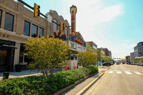 Historic theater marquee with colorful design, flanked by shops and greenery, along a city street on a sunny day.