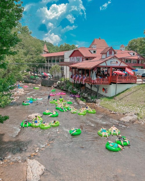 A lively river scene with people floating on green tubes, surrounded by trees and a charming building in the background.