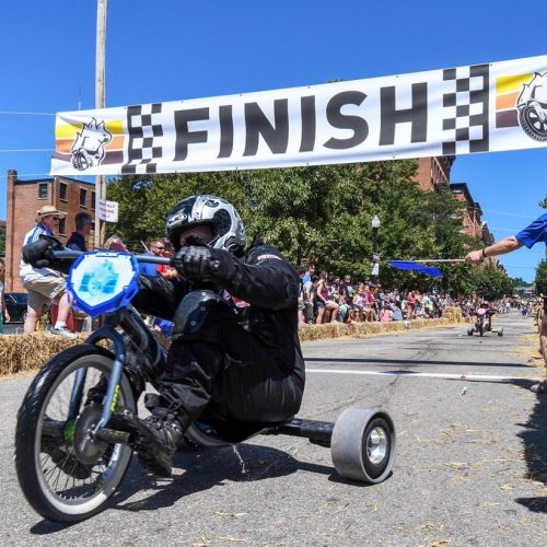 A racer in a black suit crosses the finish line on a tricycle, with a cheering crowd and a "FINISH" banner above.