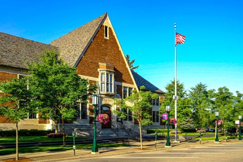 Historic brick building with a steep roof, surrounded by trees and flowers, featuring an American flag on a pole.