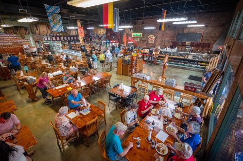 A bustling restaurant interior with wooden tables, patrons enjoying meals, and a display of food items in the background.