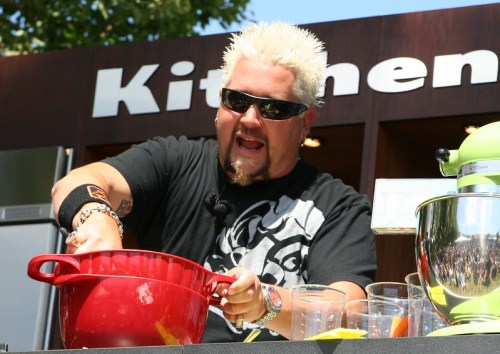 A chef with spiky blonde hair mixes ingredients in a red bowl, smiling and wearing sunglasses at a cooking demonstration.