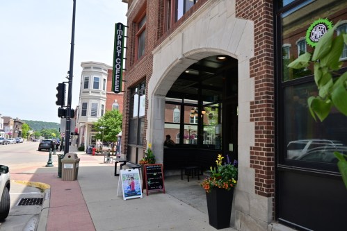 A coffee shop entrance with flowers, a sign, and a view of a street in a small town. Bright and inviting atmosphere.