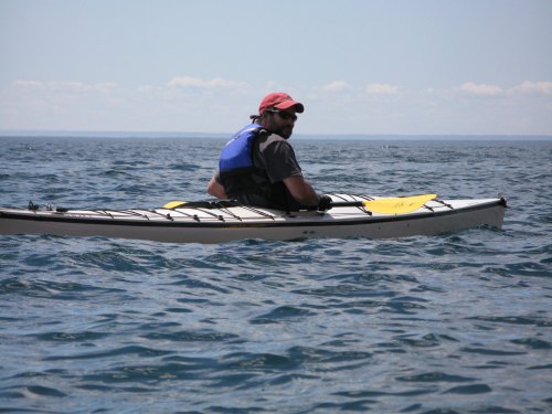 A person in a kayak wearing a red cap and life jacket, paddling on a calm blue lake under a clear sky.