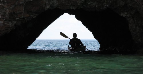 A silhouette of a person kayaking through a rocky archway, with calm waters and a clear sky in the background.