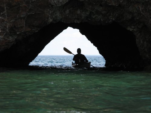 A person in a kayak paddles through a rocky archway, with calm turquoise waters and a distant horizon.