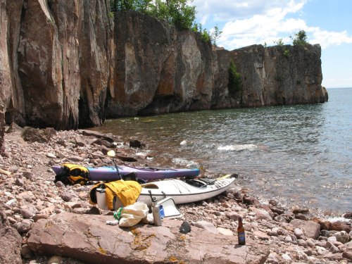 A rocky shoreline with kayaks on the beach, surrounded by cliffs and calm water under a blue sky.