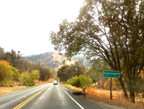 A scenic road leads to Coarsegold, framed by trees and hills in autumn colors under a bright sky.