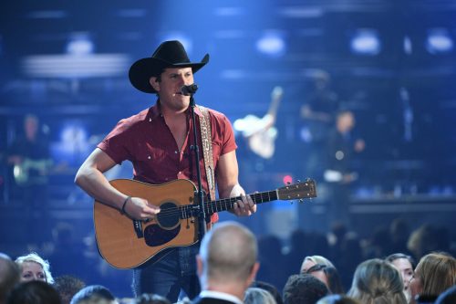 A country singer in a cowboy hat performs on stage with an acoustic guitar, surrounded by an audience.