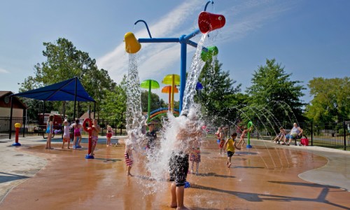 Children play in a splash pad, with colorful water features and bright sunshine in a park setting.