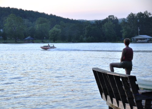 A person fishing from a dock as a speedboat passes by on a calm lake at sunset.