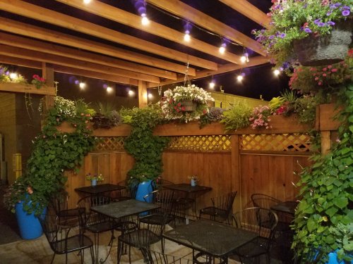 A cozy outdoor dining area with string lights, wooden beams, and lush greenery in planters.