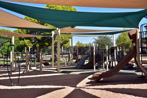 A playground with slides and climbing structures, shaded by large canopies, surrounded by trees and grass.