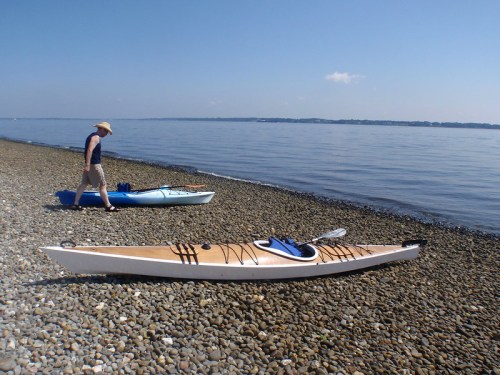 A person walks along a pebbly beach beside two kayaks on calm water under a clear blue sky.