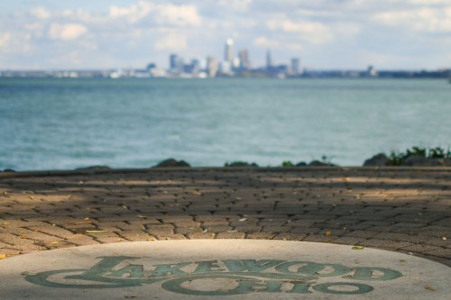 View of Lakewood sign with a city skyline in the background, overlooking a calm lake under a cloudy sky.