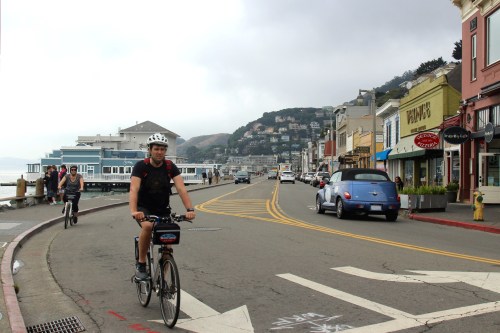 A cyclist rides along a coastal road lined with shops and buildings, with a view of the water and hills in the background.