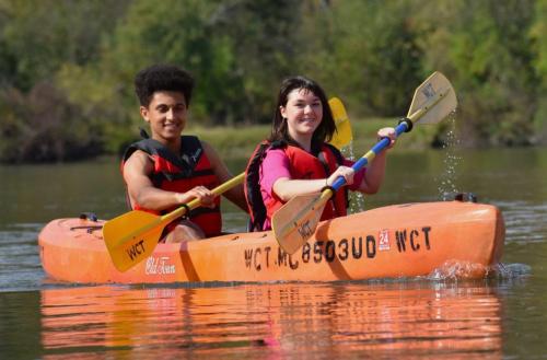 Two people in a kayak on a calm lake, smiling and paddling with vibrant green trees in the background.
