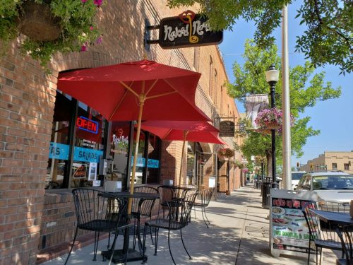 Outdoor dining area of a restaurant with red umbrellas, brick facade, and a sunny street lined with trees.