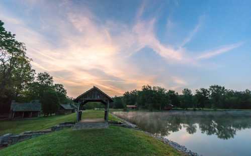 A serene lakeside scene at dawn, featuring a wooden gazebo and mist rising from the calm water under a colorful sky.