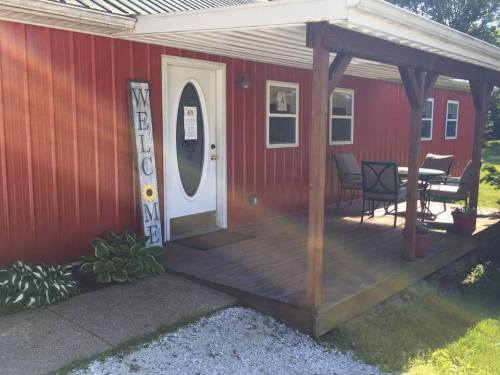 A red house with a welcoming porch, featuring a "Welcome" sign, chairs, and potted plants. Sunlight shines on the scene.