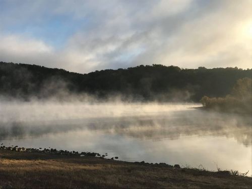 A serene lake shrouded in morning mist, surrounded by hills under a cloudy sky.