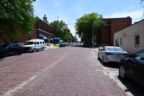 A brick-paved street lined with parked cars and trees, with buildings and a clock tower in the background under a clear sky.