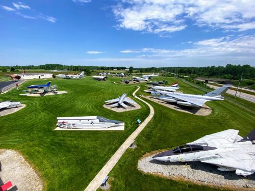 Aerial view of a grassy area displaying various military aircraft and jets in a museum setting under a blue sky.