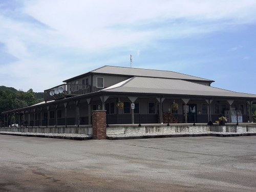 A large, two-story building with a gray roof and porch, surrounded by a paved area and trees in the background.