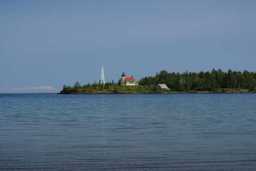 A serene island with a lighthouse and a small building, surrounded by calm water and lush greenery under a clear sky.