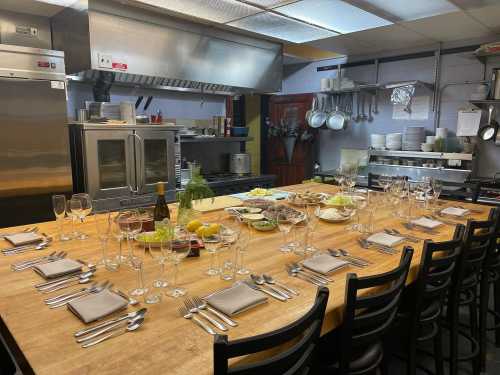 A spacious kitchen with a large wooden table set for a meal, featuring glassware, utensils, and various food items.