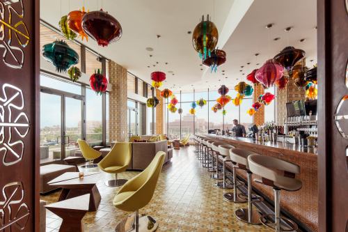 A stylish bar with colorful hanging lanterns, modern seating, and large windows offering a city view.