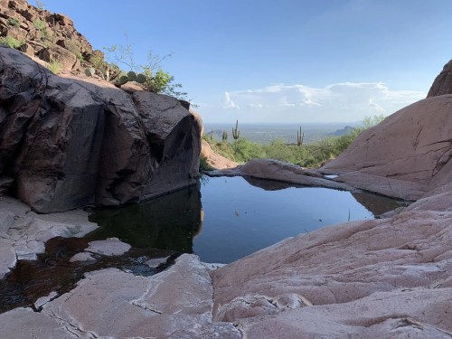A serene natural pool surrounded by rocky terrain and cacti, with a distant view of the landscape under a clear sky.