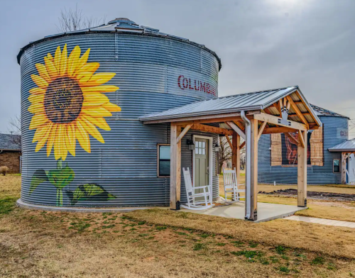 A metal silo with a large sunflower mural, featuring a porch with rocking chairs in front.