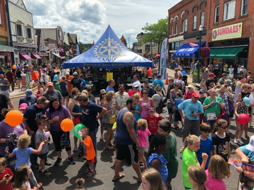 A lively street festival with a large crowd, colorful balloons, and booths, set in a charming town.